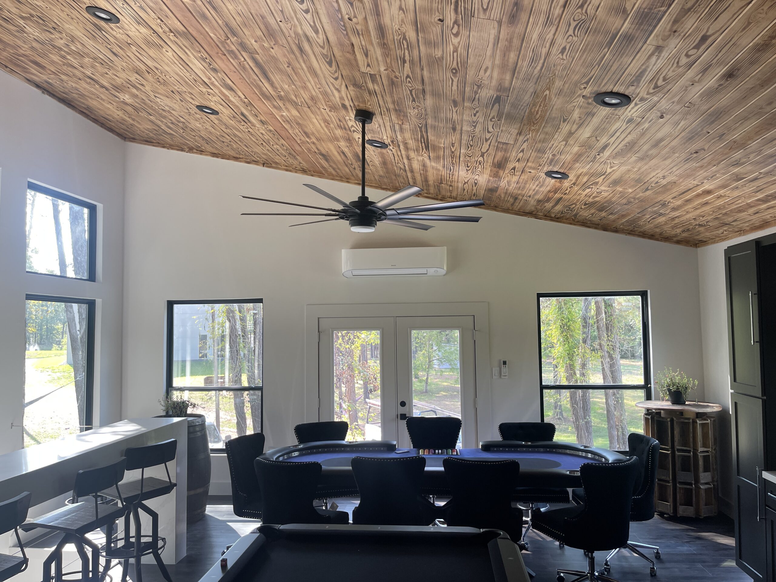 Room with a wooden ceiling, large windows, a ceiling fan, poker table with chairs, bar stools, and a pool table in the foreground. Natural light fills the space.