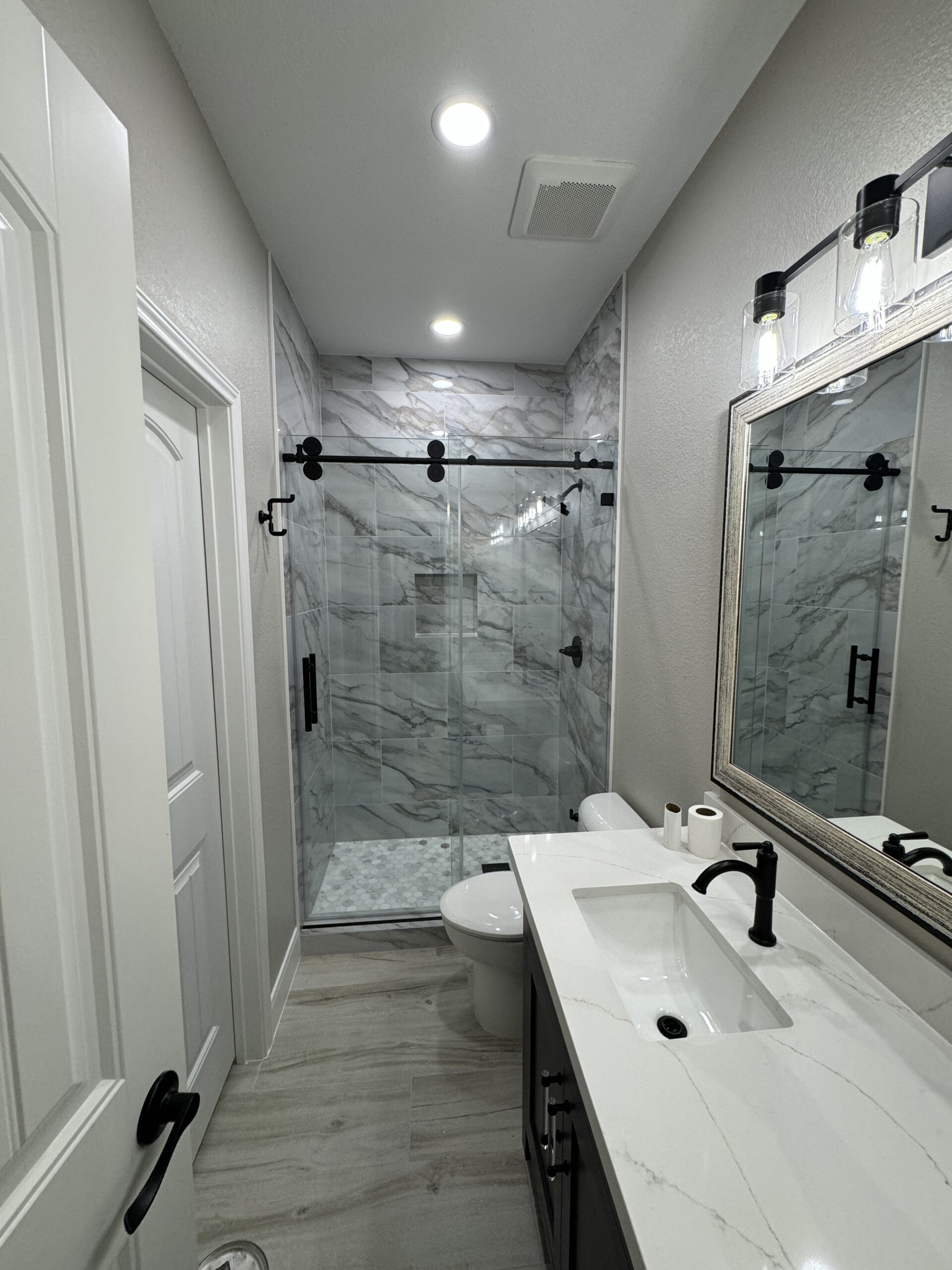 Modern bathroom with a glass-enclosed marble shower, white countertop sink, toilet, light gray walls, and ceiling lights.