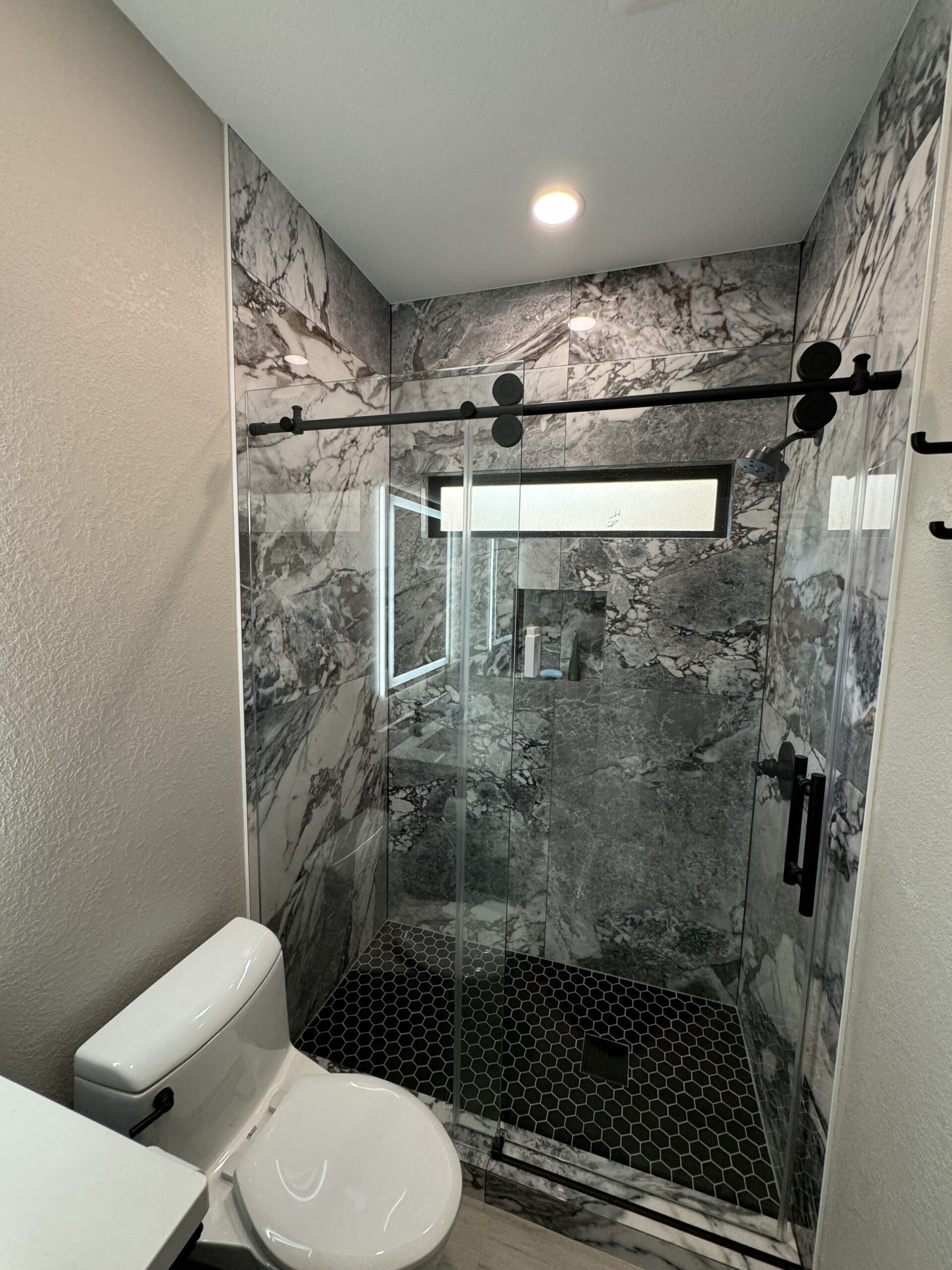 Modern bathroom with a glass-enclosed shower featuring gray marble walls, a black hexagonal tile floor, a window, and a white toilet in the foreground.