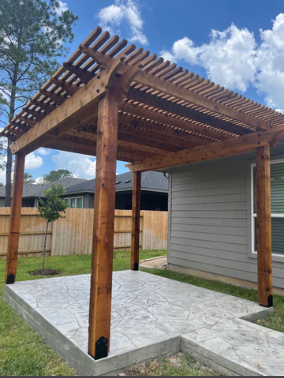A wooden pergola stands on a stamped concrete patio beside a house, with a wooden fence and trees in the background under a partly cloudy sky.