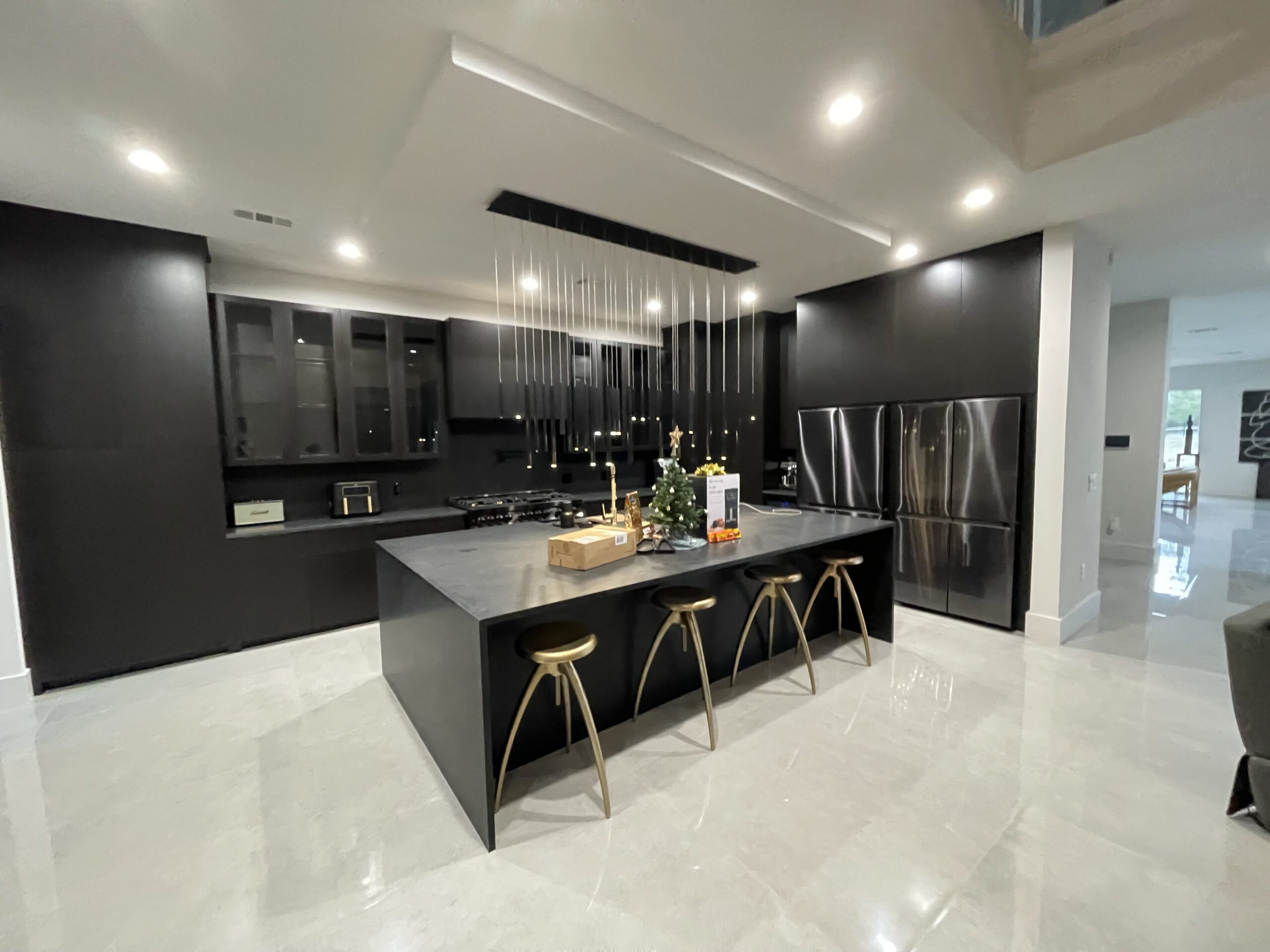 Modern kitchen with black cabinetry and countertops, three gold stools at an island, and stainless steel appliances; small holiday decorations on the island.