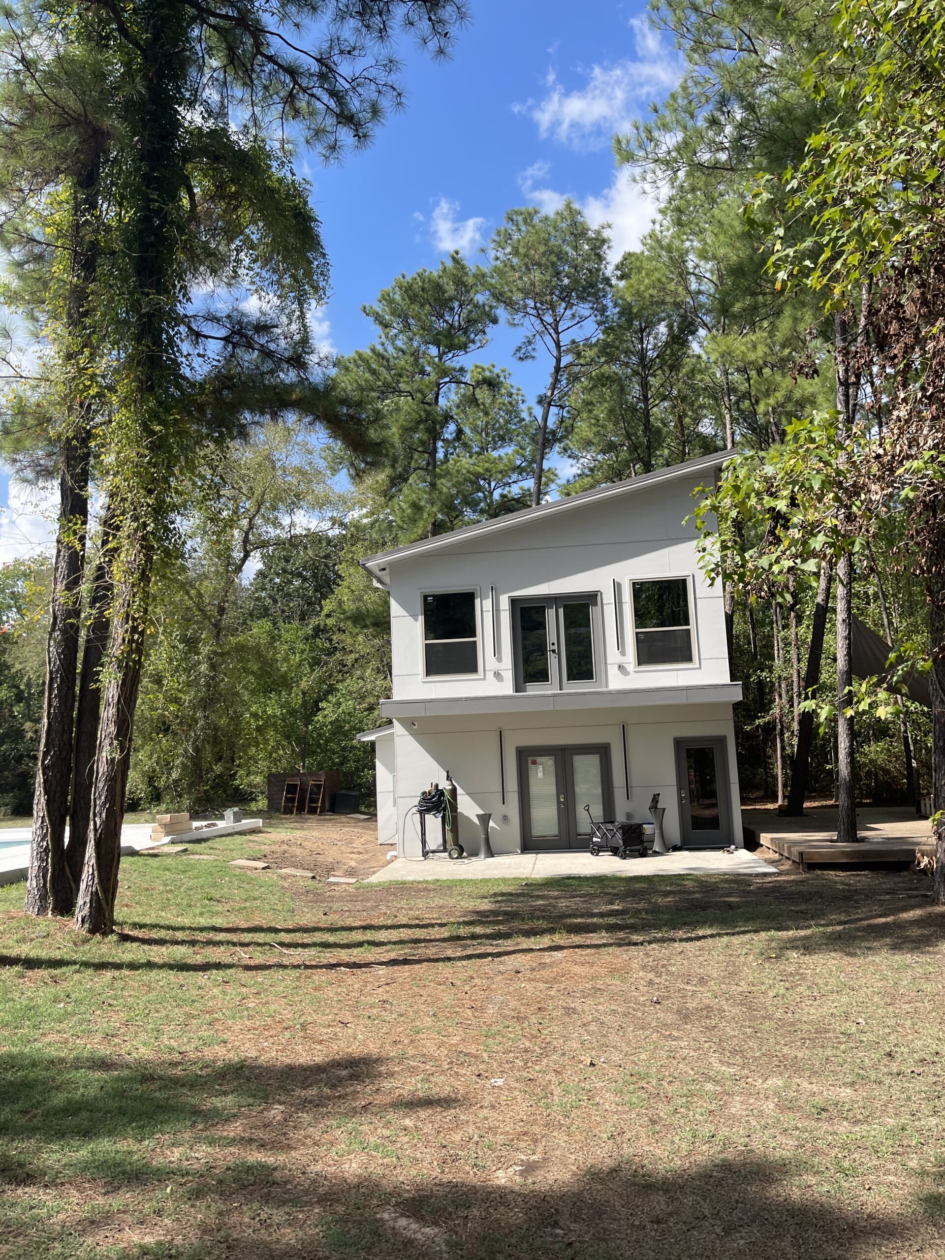 A modern two-story gray house with large windows sits in a grassy, wooded area under a blue sky with scattered clouds.